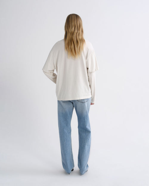 A woman with long blonde hair is shown from behind wearing an oversized off-white long-sleeve top and light-wash blue jeans against a plain light gray studio background.