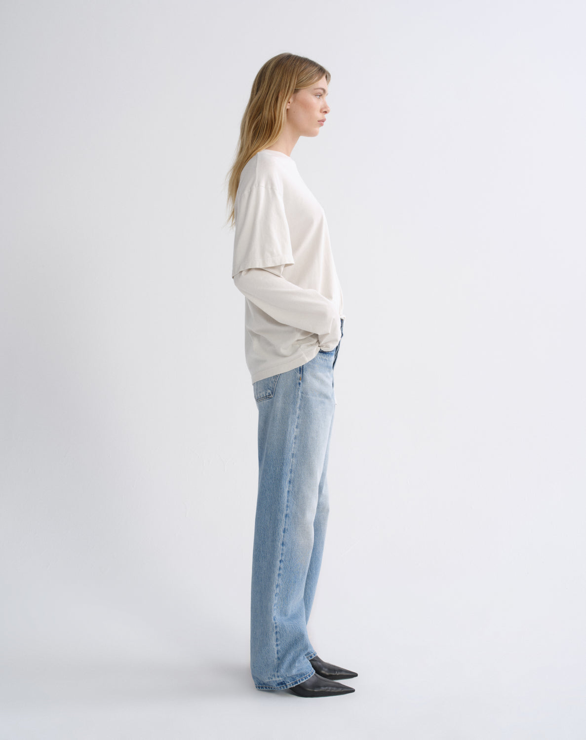 A woman stands in profile wearing a cream long-sleeve top, light blue wide-leg jeans, and black pointed flats against a plain light gray studio background.