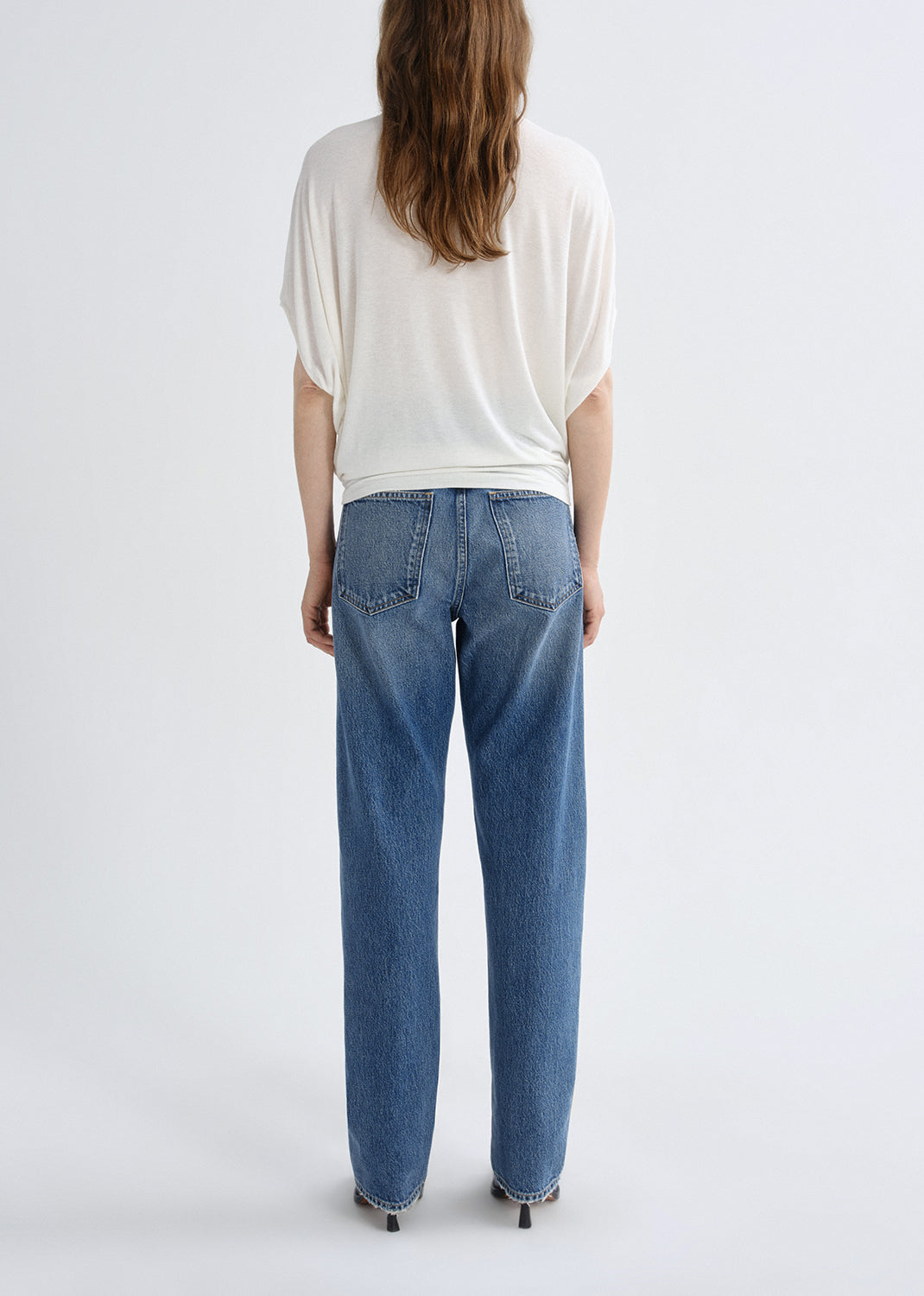 Person wearing a white short sleeve crewneck tee with light wash straight leg jeans viewed from the back on a white background