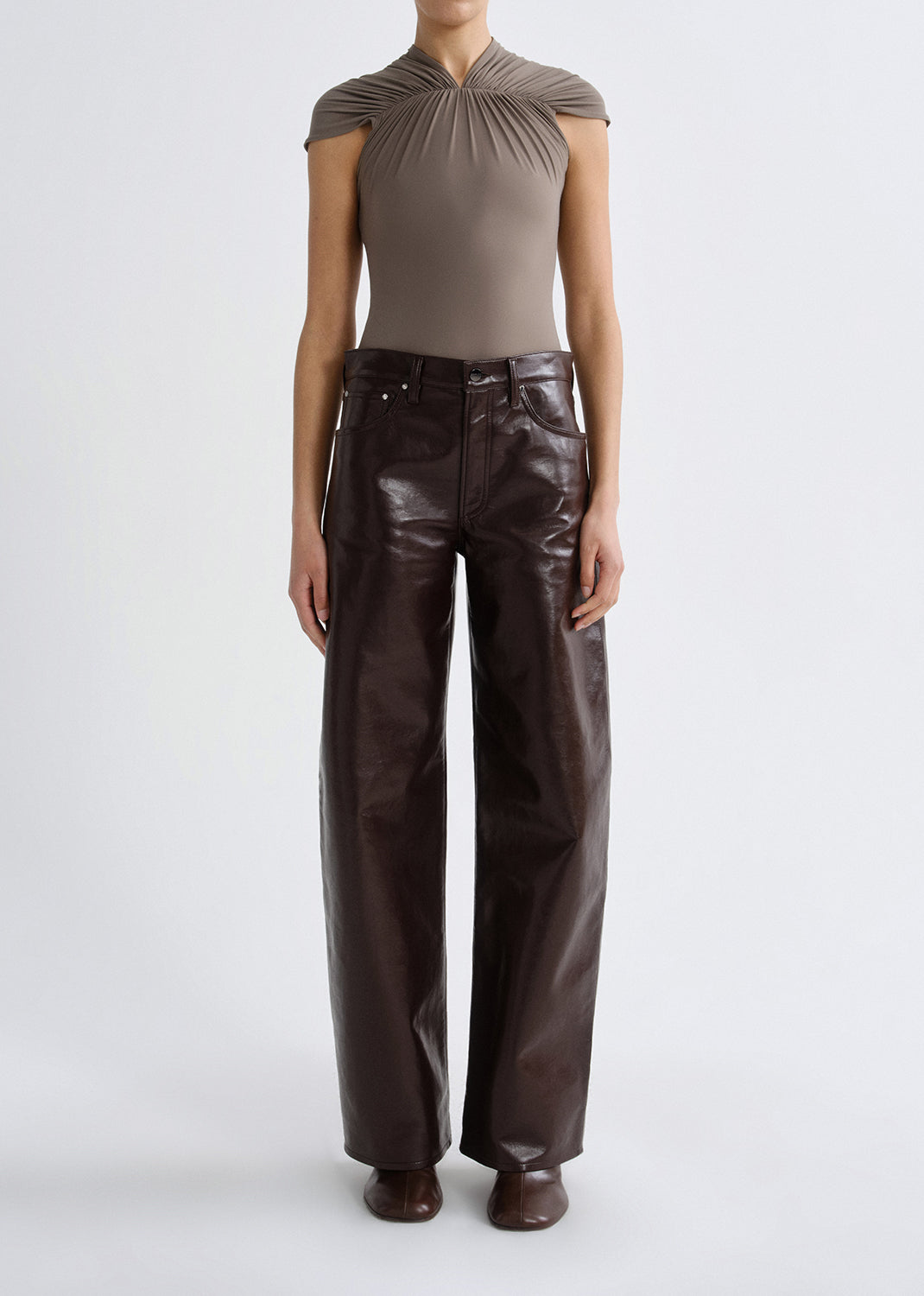 Person wearing a taupe cap sleeve draped top with dark brown leather pants on a grey background.