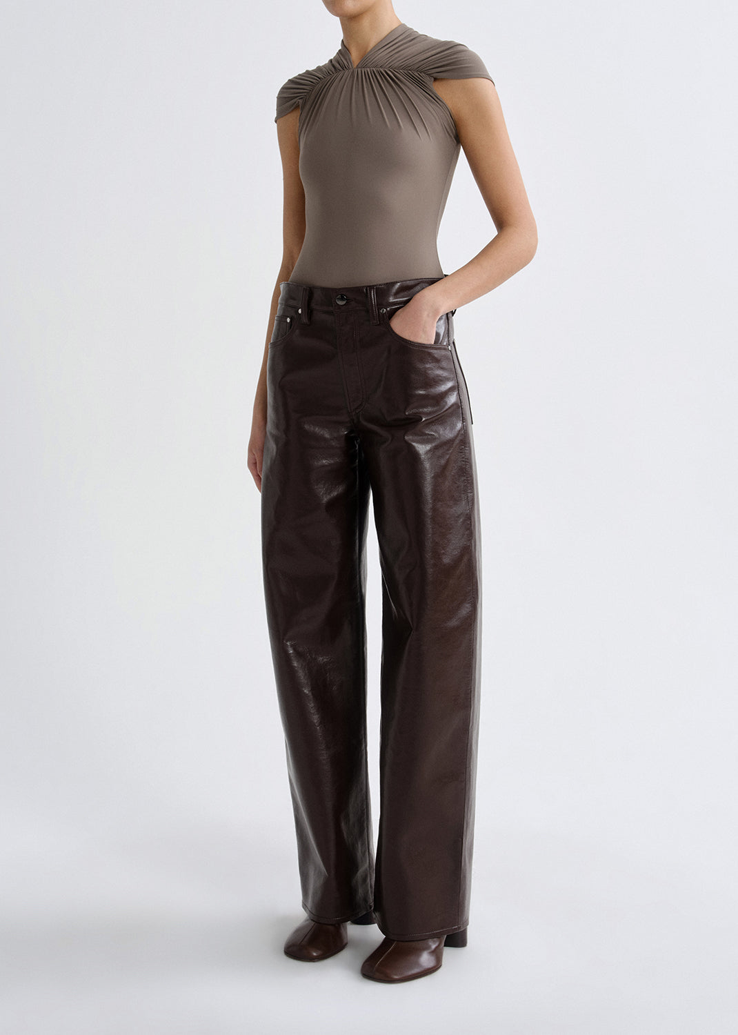 Person wearing a taupe cap sleeve draped top with dark brown leather pants on a grey background.