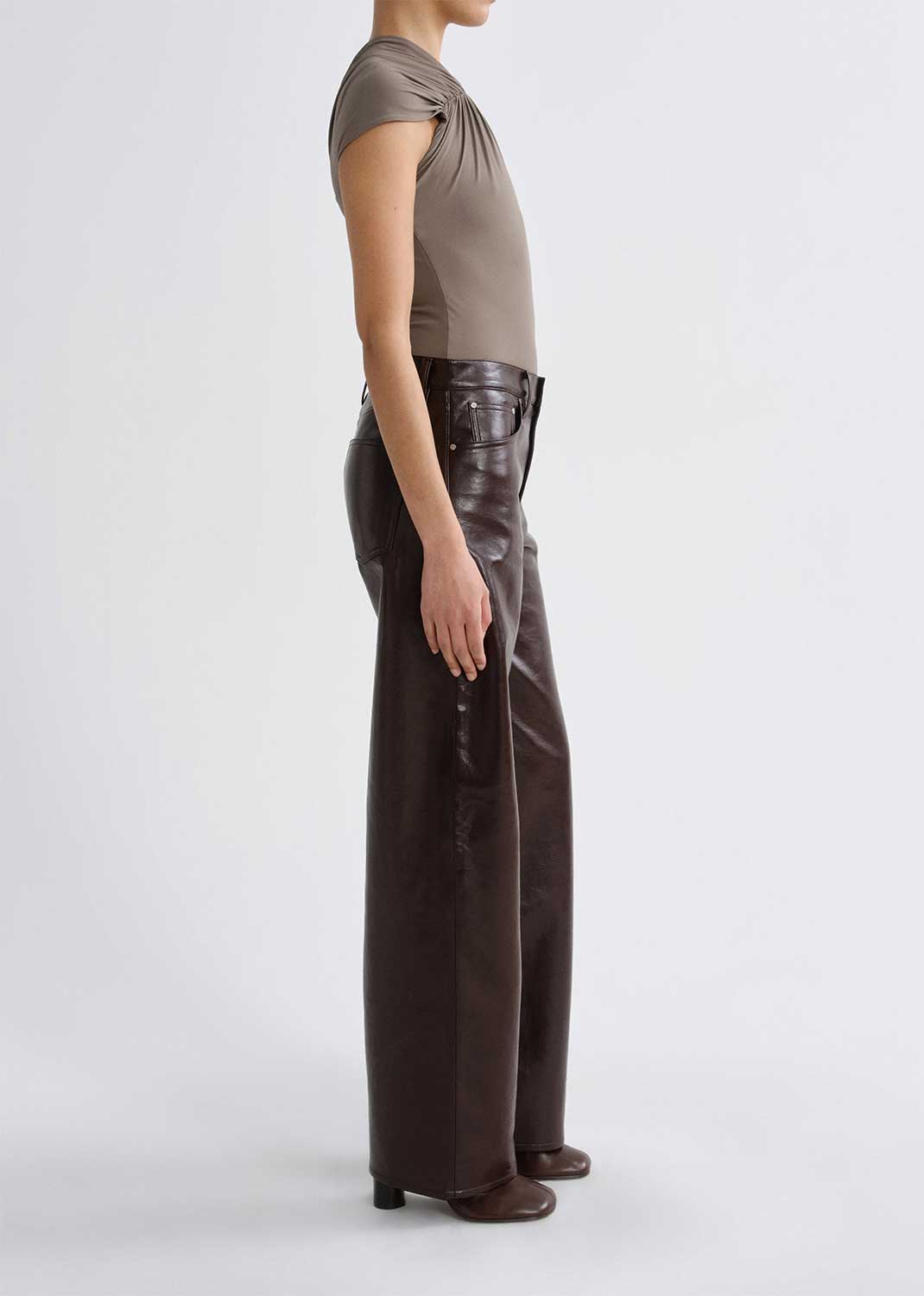 Person wearing a taupe cap sleeve draped top with dark brown leather pants viewed from the side on a grey background.
