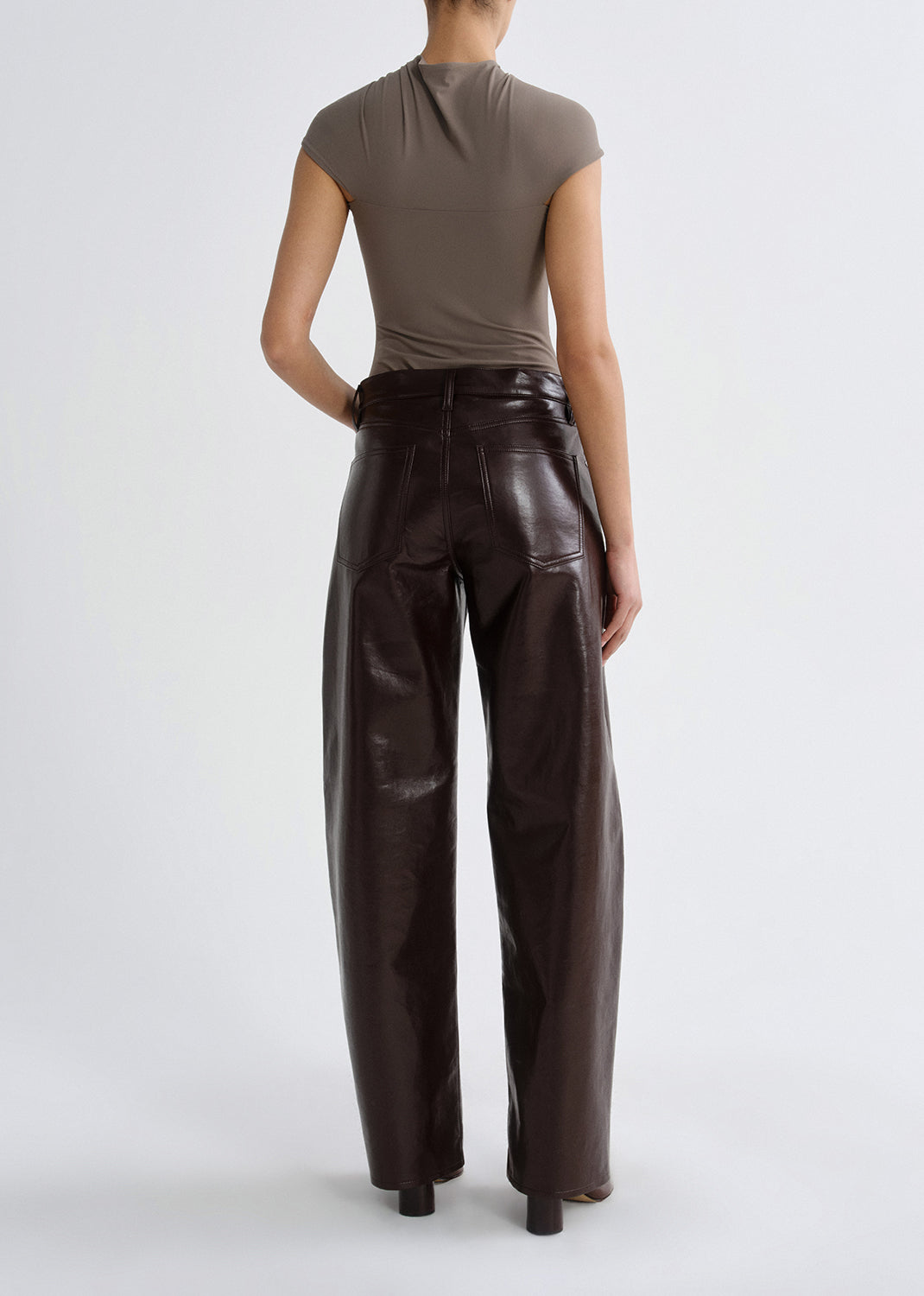 Person wearing a taupe cap sleeve draped top with dark brown leather pants viewed from the back on a grey background.