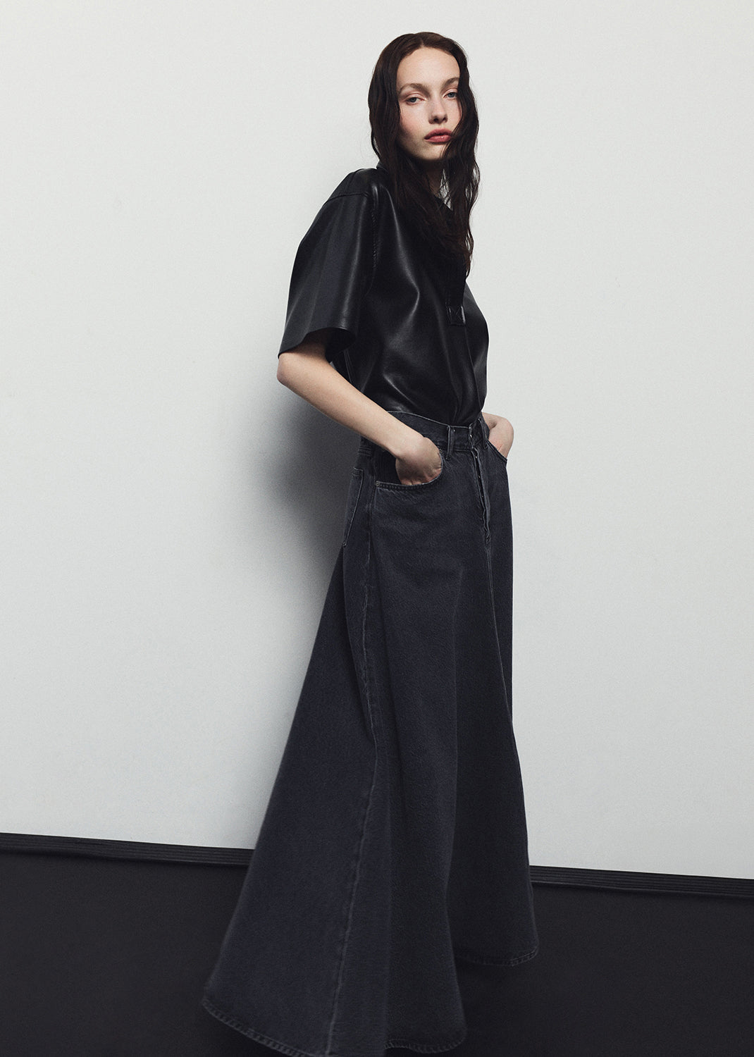 Person wearing a black outfit with a long black skirt against a white wall.