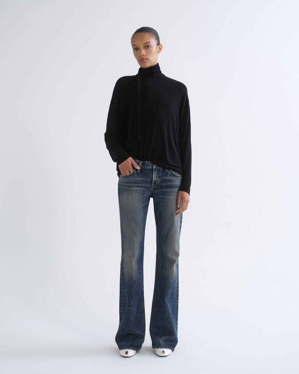 A model stands against a light gray studio background wearing a black long-sleeve mock-neck top, blue flare jeans, and white pointed shoes.