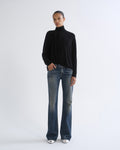A model stands against a light gray studio background wearing a black long-sleeve mock-neck top, blue flare jeans, and white pointed shoes.