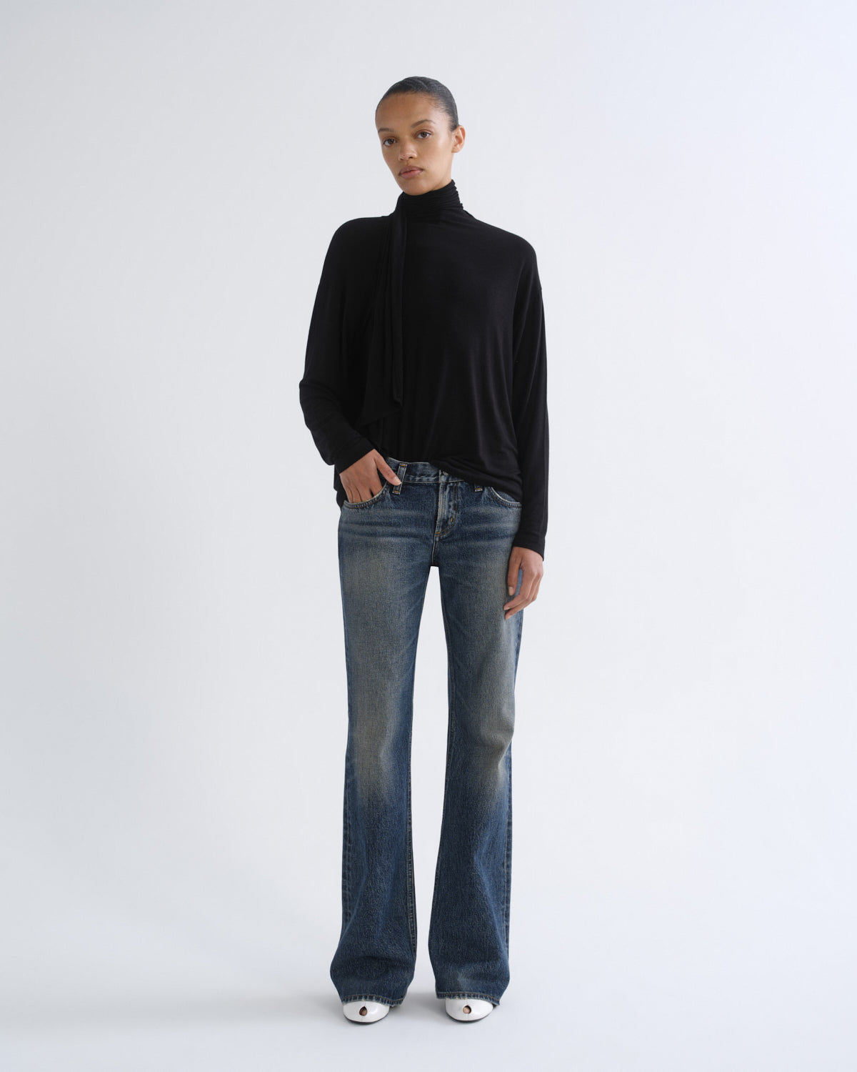 A model stands against a light gray studio background wearing a black long-sleeve mock-neck top, blue flare jeans, and white pointed shoes.