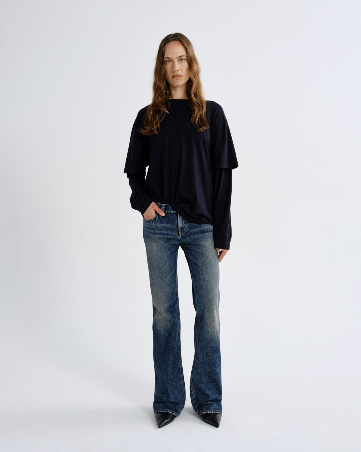 A woman stands against a light gray studio background wearing a loose black long-sleeve top, blue flare jeans, and black pointed shoes.