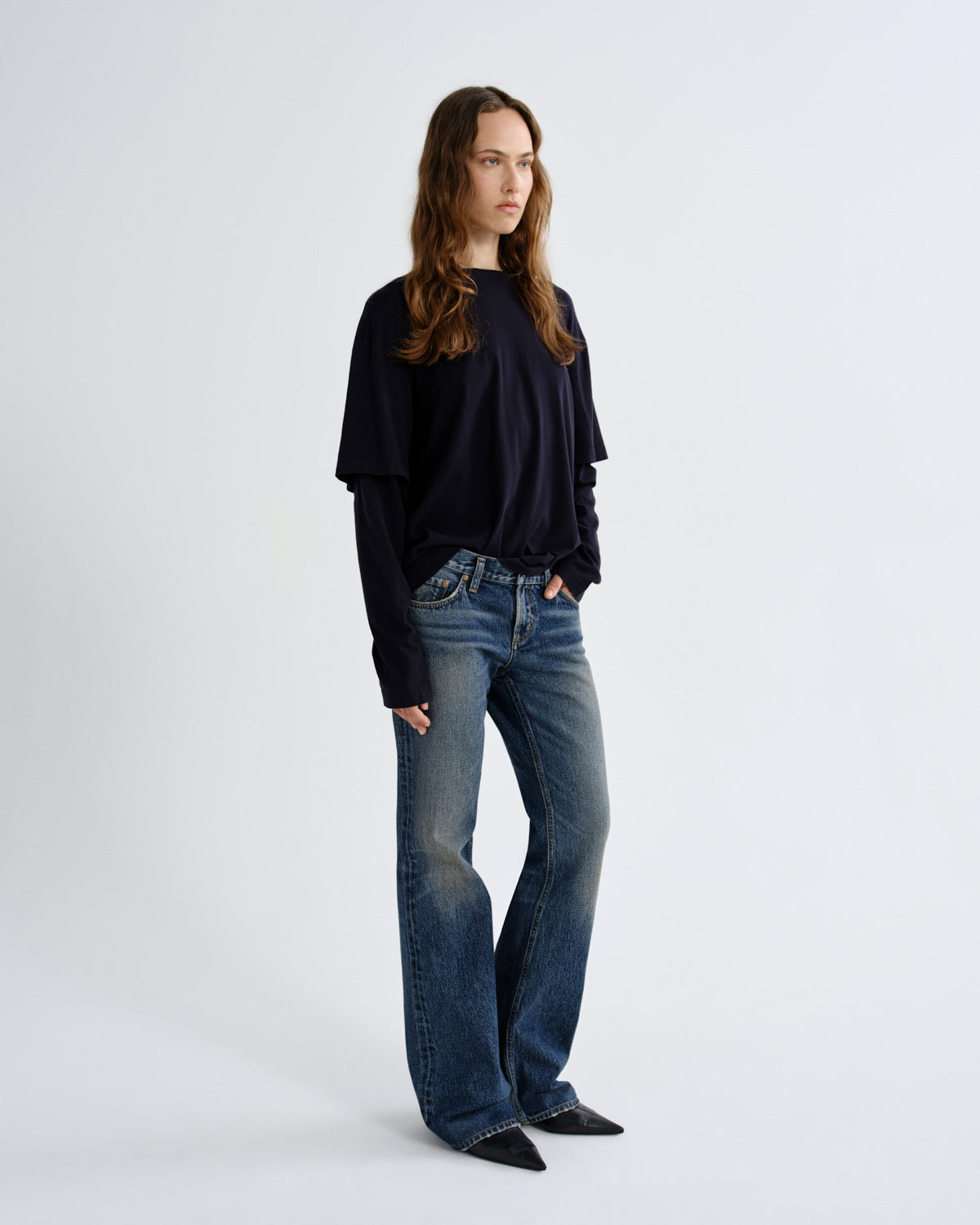 A woman modeling a dark navy long-sleeve top with layered ruffle details on the sleeves and blue flare jeans, standing against a light gray studio background.