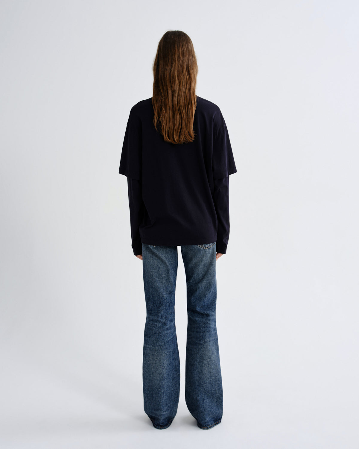 A woman viewed from behind wearing a loose black long-sleeve top and blue flared jeans against a plain light gray studio background.