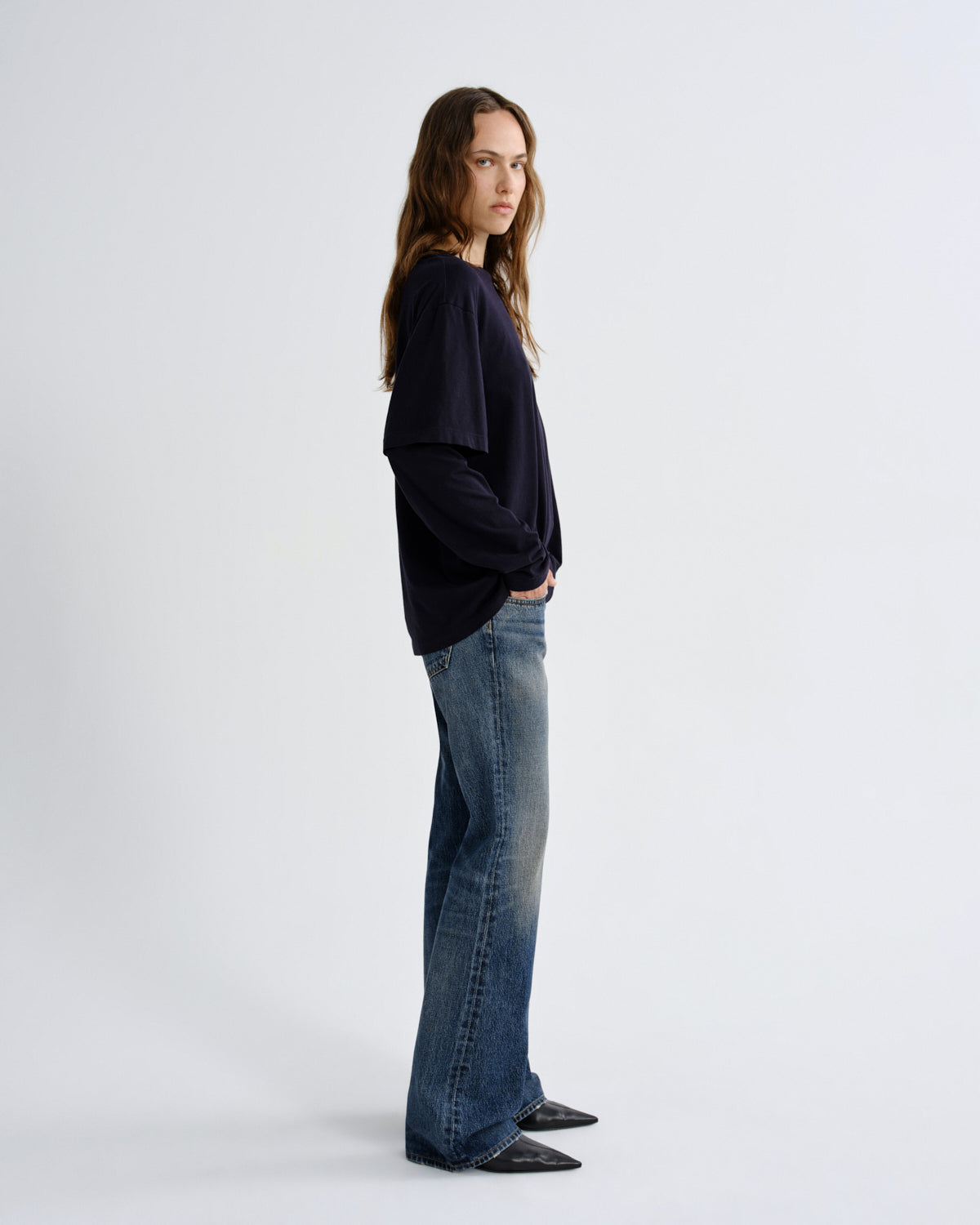 A woman stands in profile wearing a loose dark navy long-sleeve top, blue wide-leg jeans, and black pointed flats against a clean light gray studio background.