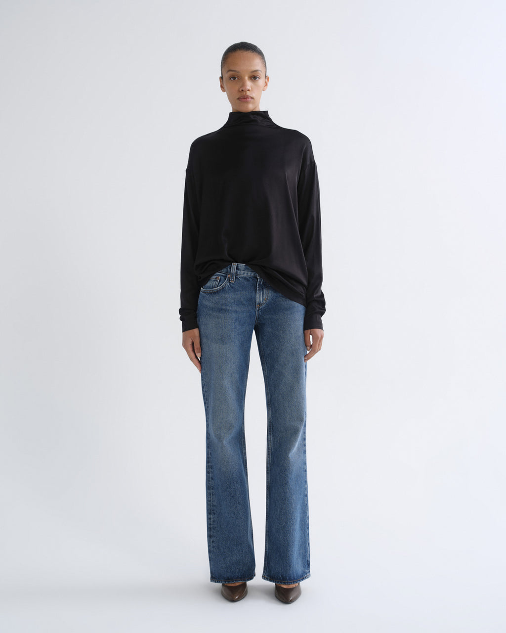 A model stands against a light gray studio background wearing a black long-sleeve mock-neck top and blue straight-leg jeans.