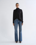 A model stands against a light gray studio background wearing a black long-sleeve mock-neck top and blue straight-leg jeans.
