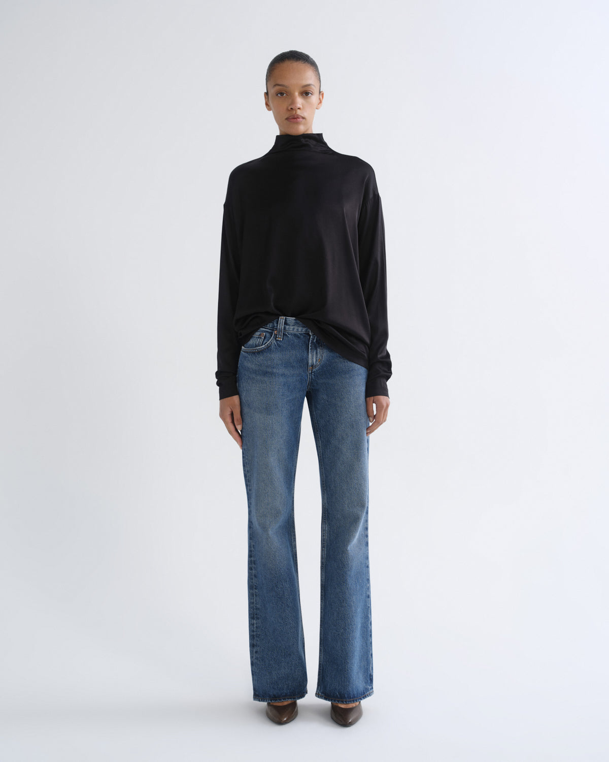 A model stands against a light gray studio background wearing a black long-sleeve mock-neck top and blue straight-leg jeans.