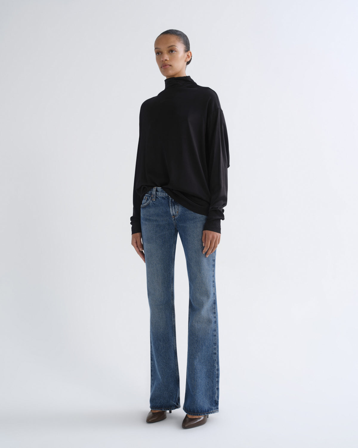 A model stands against a light gray studio background wearing a black long-sleeve turtleneck top, blue straight-leg jeans, and brown pointed shoes.
