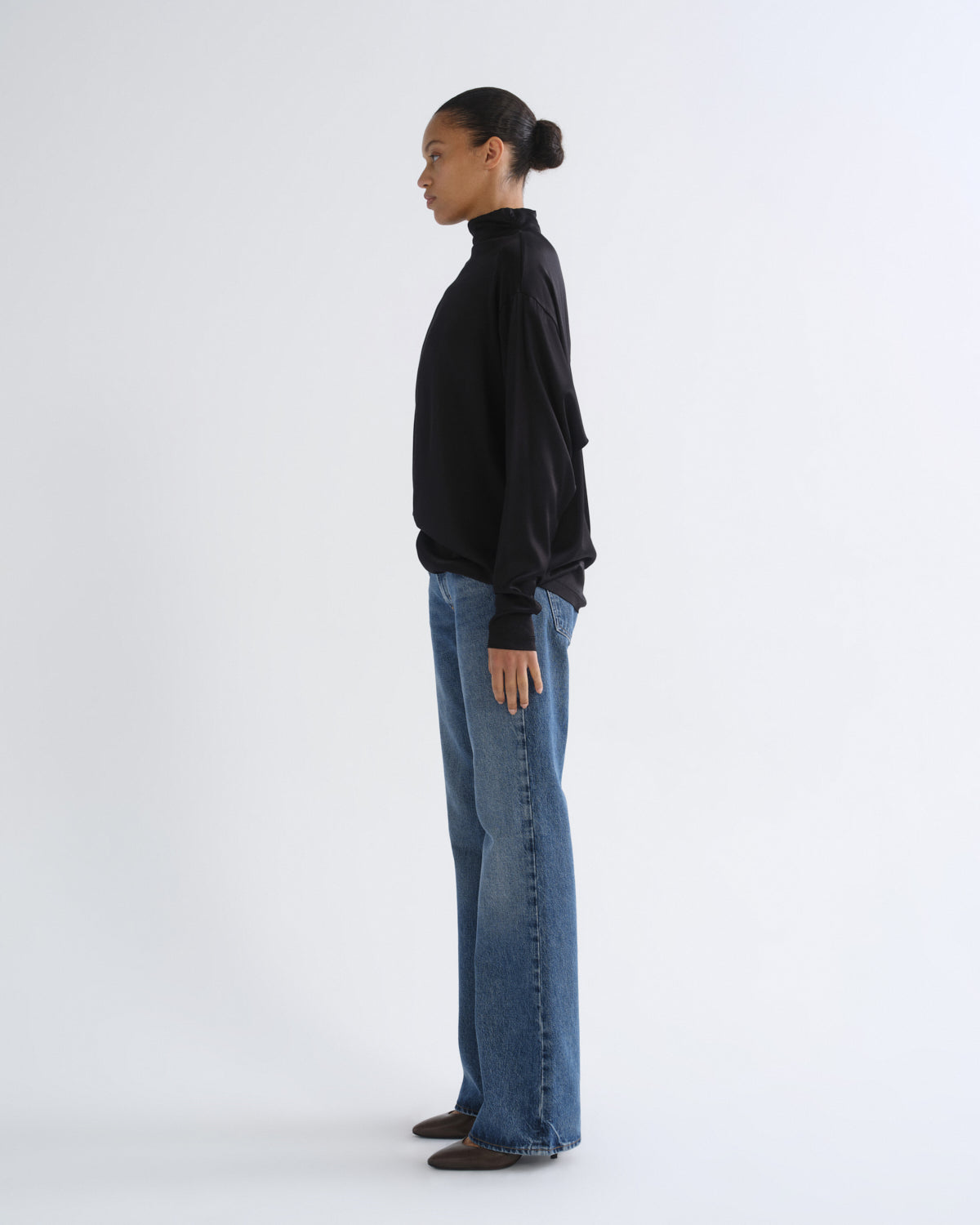 A model shown in profile wearing a black long-sleeve mock neck top, blue wide-leg jeans, and dark brown shoes, standing against a plain light gray studio background.