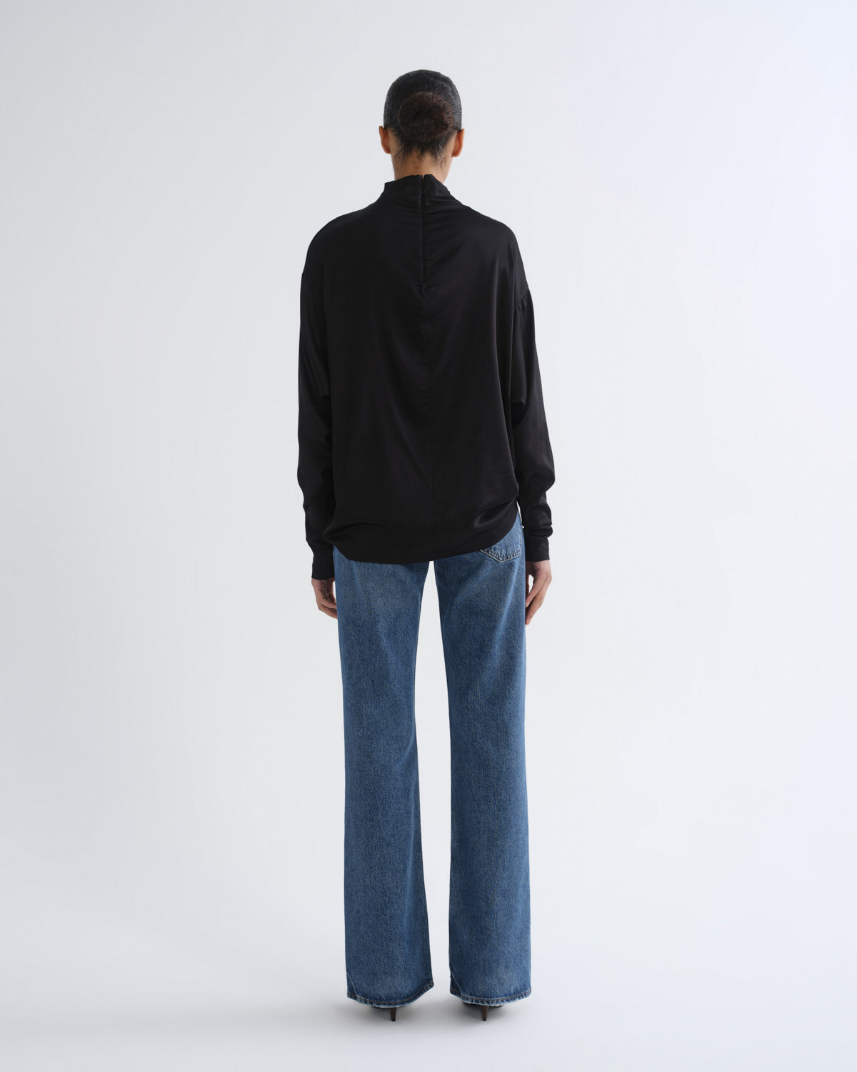 A model viewed from the back wearing a black long-sleeve mock-neck top and wide-leg blue jeans against a plain white studio background.