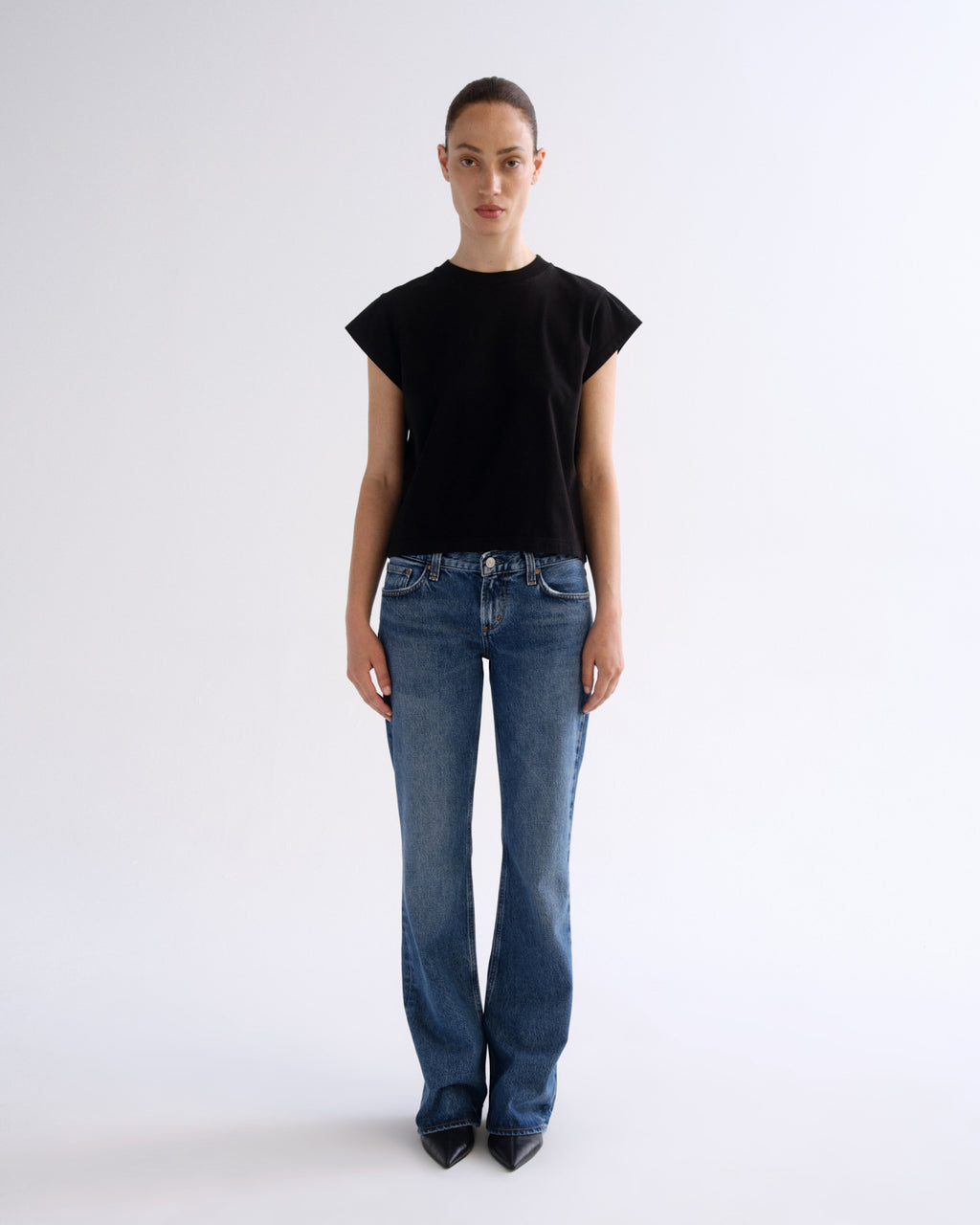 A model stands against a plain light gray studio background wearing a black short-sleeve crewneck top, blue jeans, and black pointed shoes.