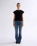 A model stands against a plain light gray studio background wearing a black short-sleeve crewneck top, blue jeans, and black pointed shoes.