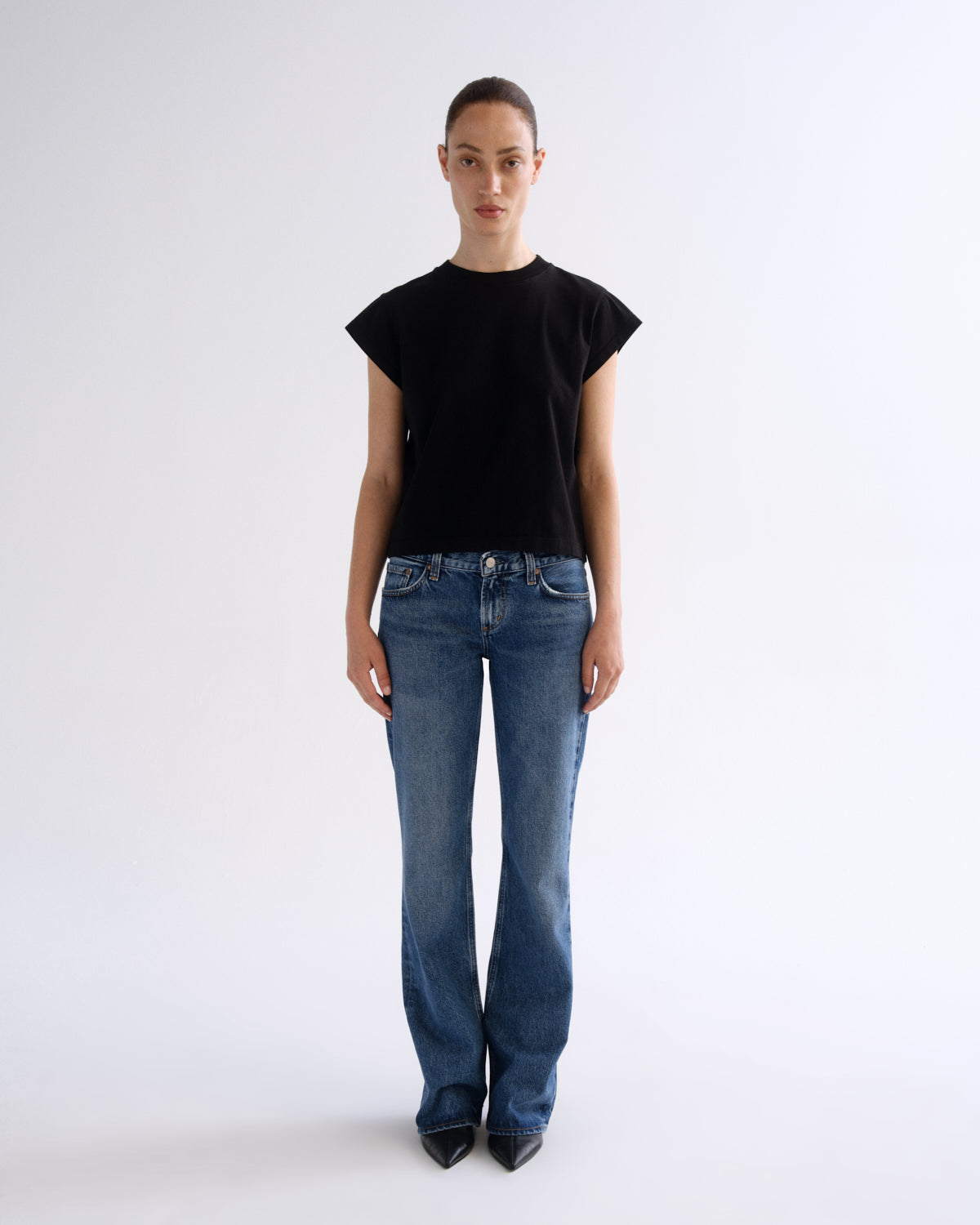 A model stands against a plain light gray studio background wearing a black short-sleeve crewneck top, blue jeans, and black pointed shoes.