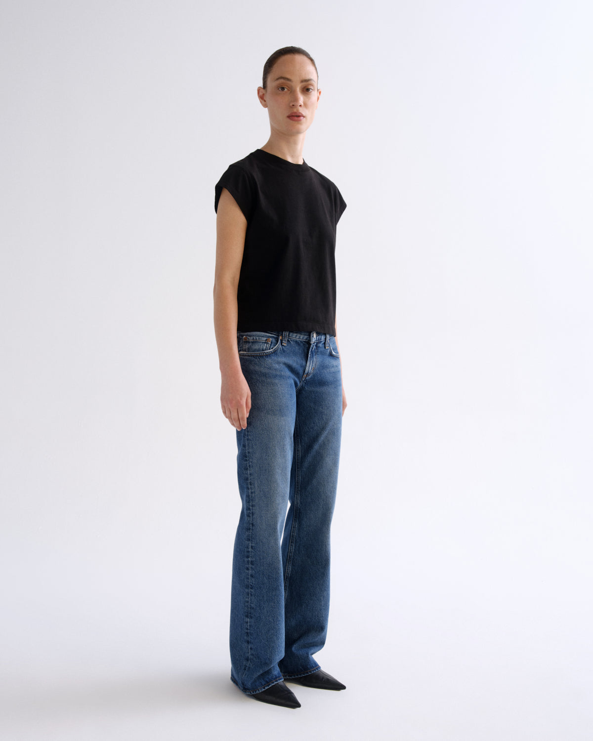 A model stands in profile wearing a black short-sleeve crewneck t-shirt and blue straight-leg jeans against a plain light gray studio background.