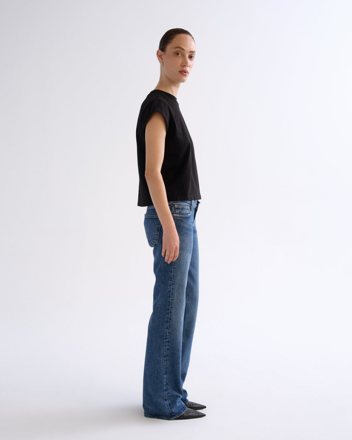 A woman stands in profile wearing a black sleeveless cropped top, blue wide-leg jeans, and dark pointed flats against a clean light gray studio background.