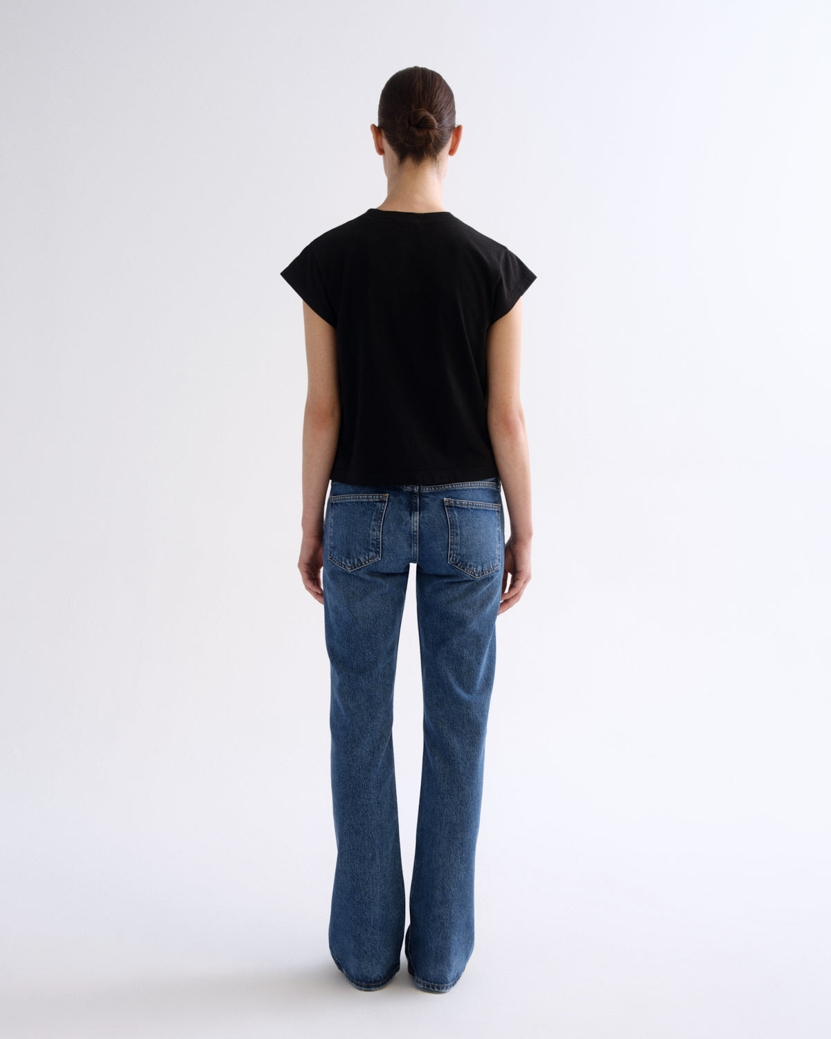 A model viewed from the back wearing a black short-sleeve t-shirt and blue flare jeans against a plain white studio background.
