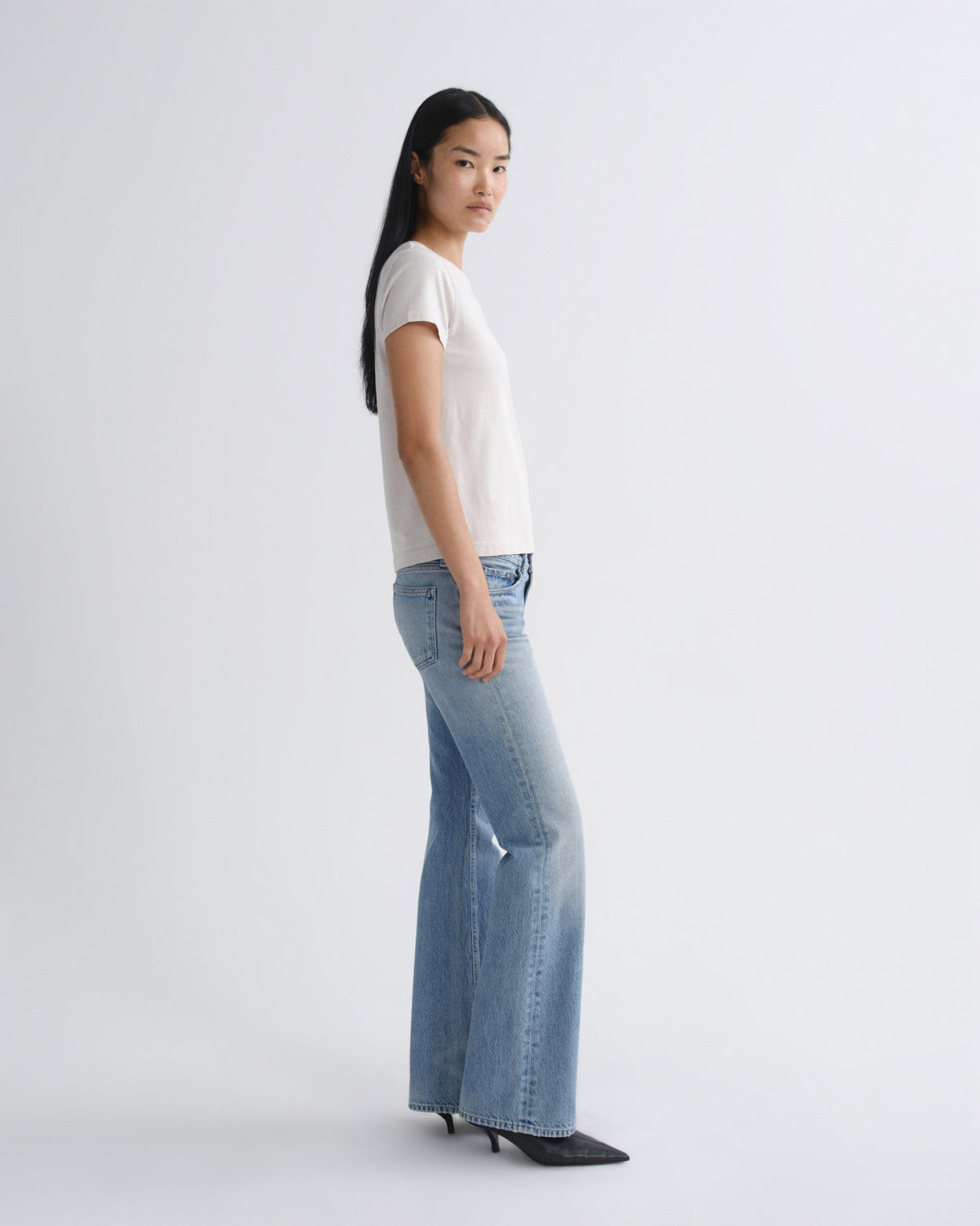 A woman stands in profile wearing a white short-sleeve top, light-wash blue flared jeans, and black pointed shoes against a clean light gray studio background.