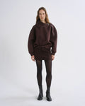 A model stands against a grey background wearing a dark brown asymmetrical zip-up sweatshirt, a matching dark brown mini skirt, black tights, and black ankle boots.