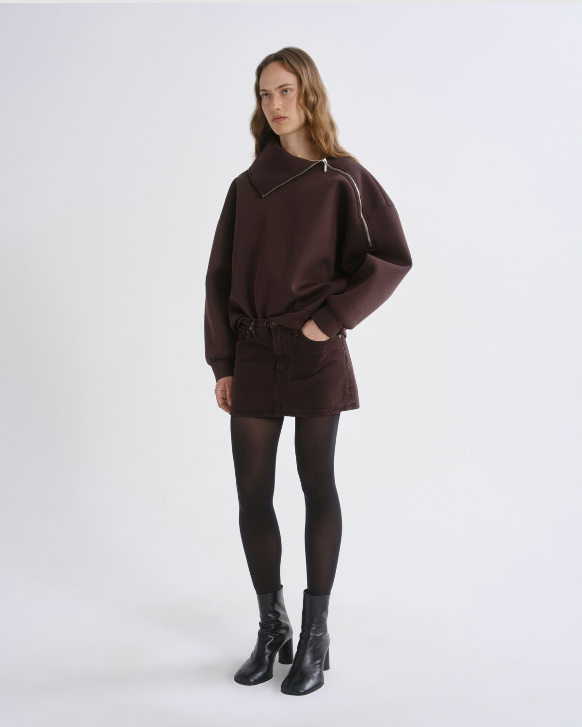 A model stands against a grey background wearing a dark brown oversized sweater dress with silver zipper details, black tights, and black ankle boots.