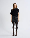 A model stands against a light gray studio background wearing a black short-sleeve top with draped shoulders, a black faux-leather mini skirt, black tights, and dark brown ankle boots.