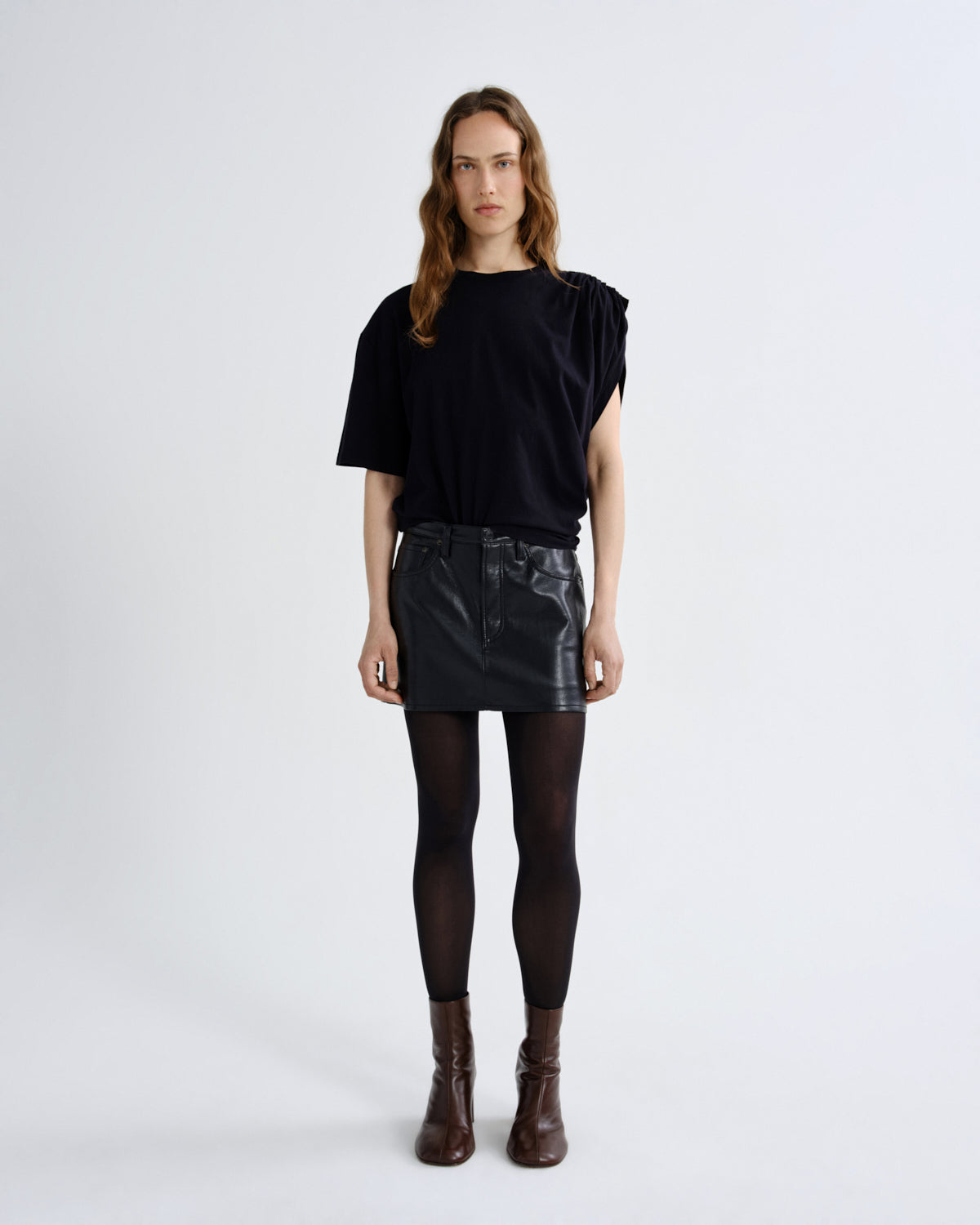 A model stands against a light gray studio background wearing a black short-sleeve top with draped shoulders, a black faux-leather mini skirt, black tights, and dark brown ankle boots.