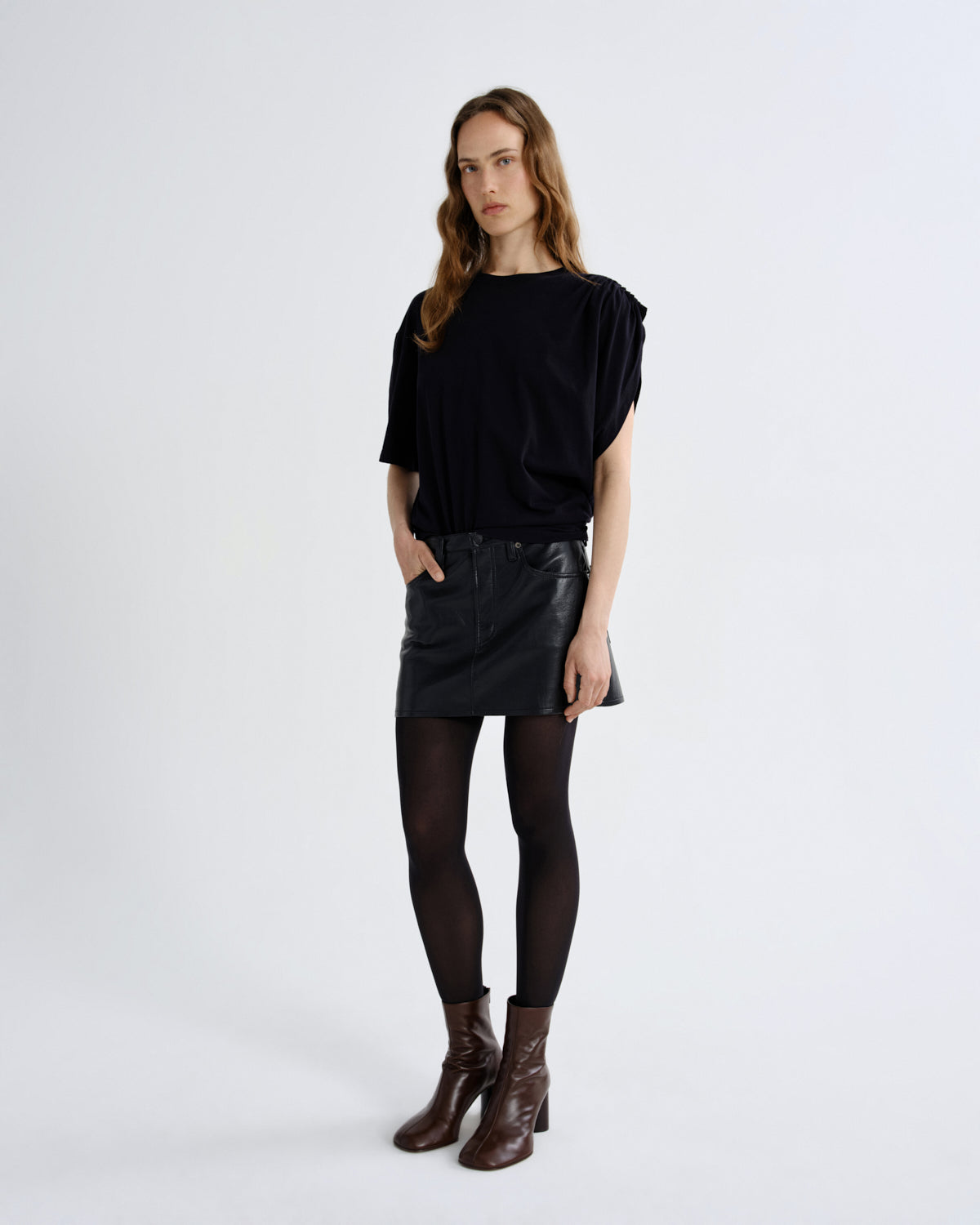 A woman modeling a black short-sleeve top, a black faux-leather mini skirt, black tights, and brown ankle boots against a plain white studio background.
