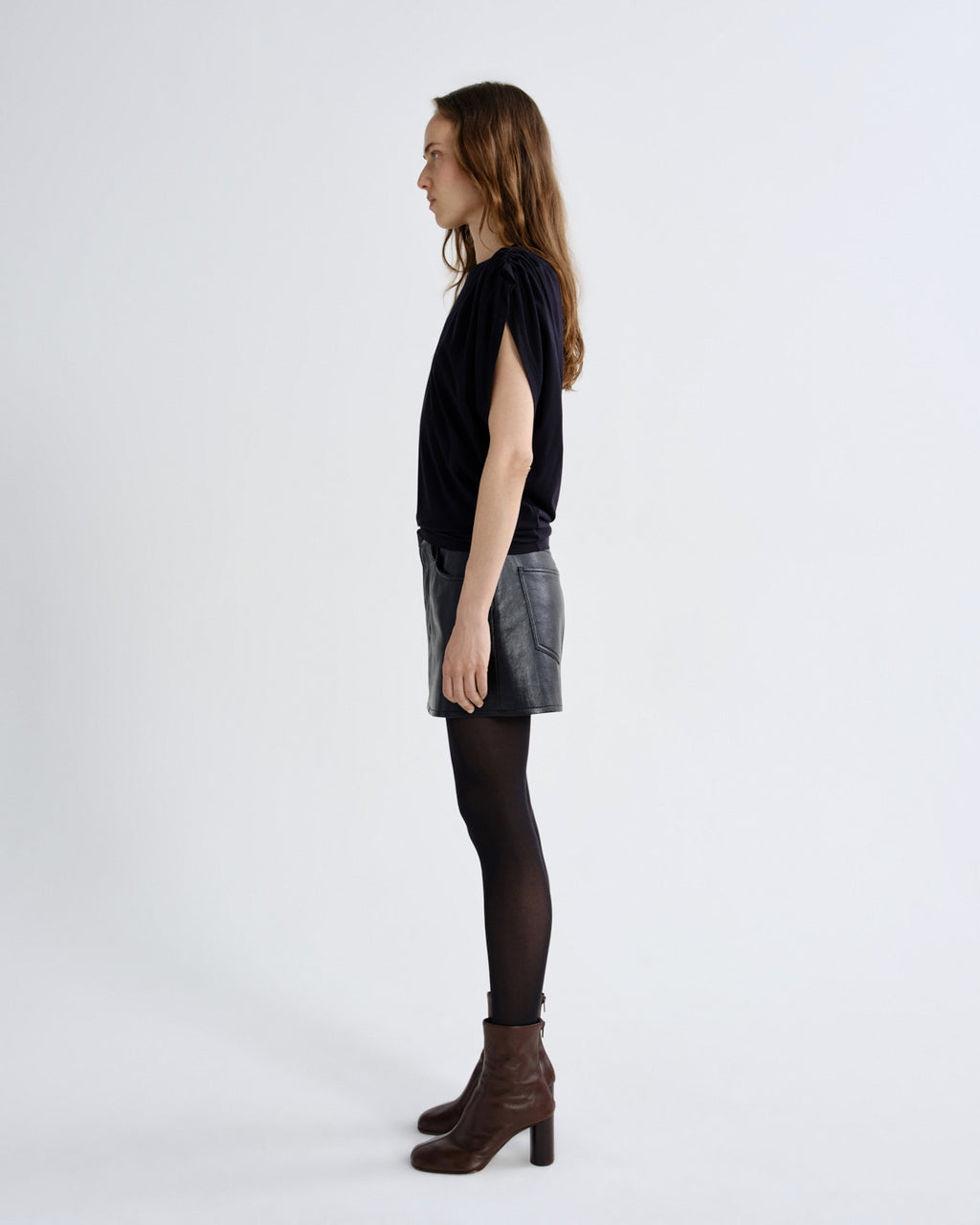 A woman stands in profile wearing a black short-sleeve top, a gray denim mini skirt, black tights, and brown heeled ankle boots against a plain light gray studio background.