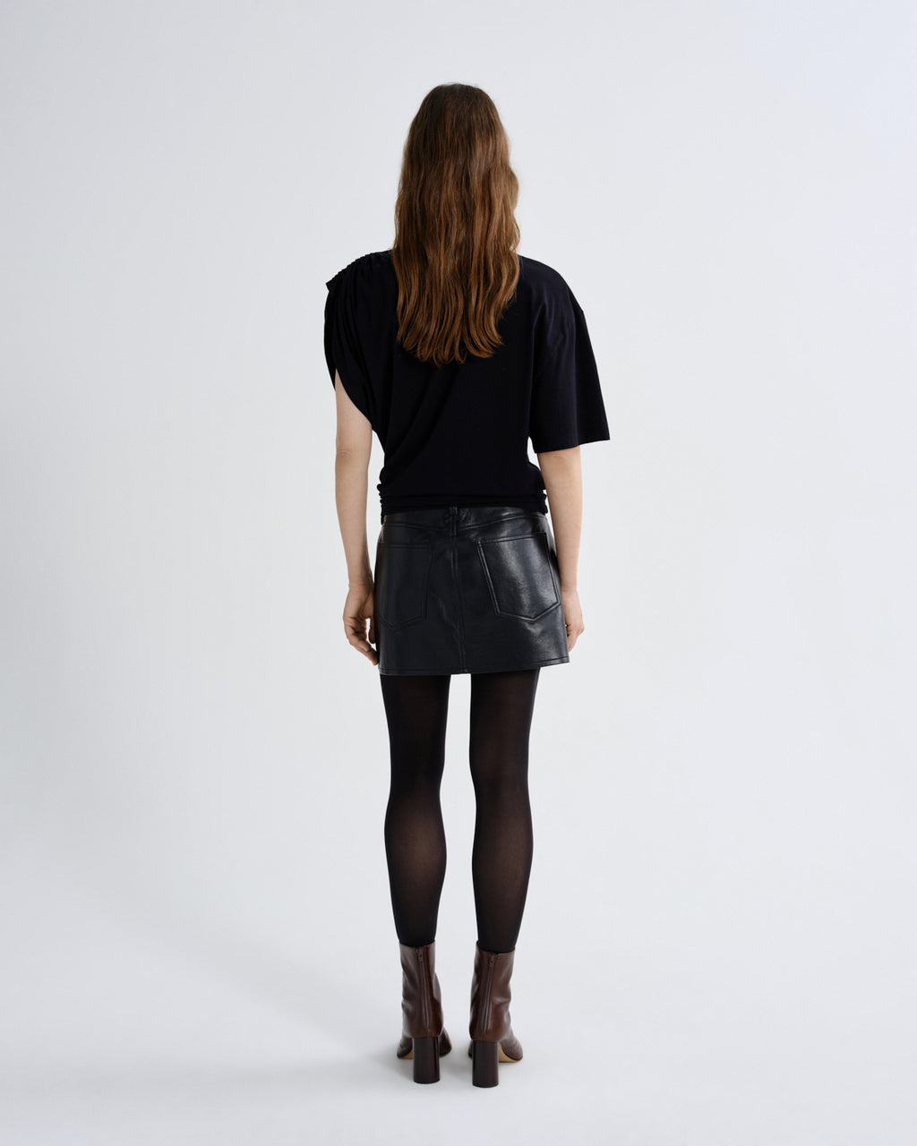 A model viewed from behind wearing a black short-sleeve top, a black faux-leather mini skirt, black tights, and brown heeled ankle boots against a plain light gray studio background.