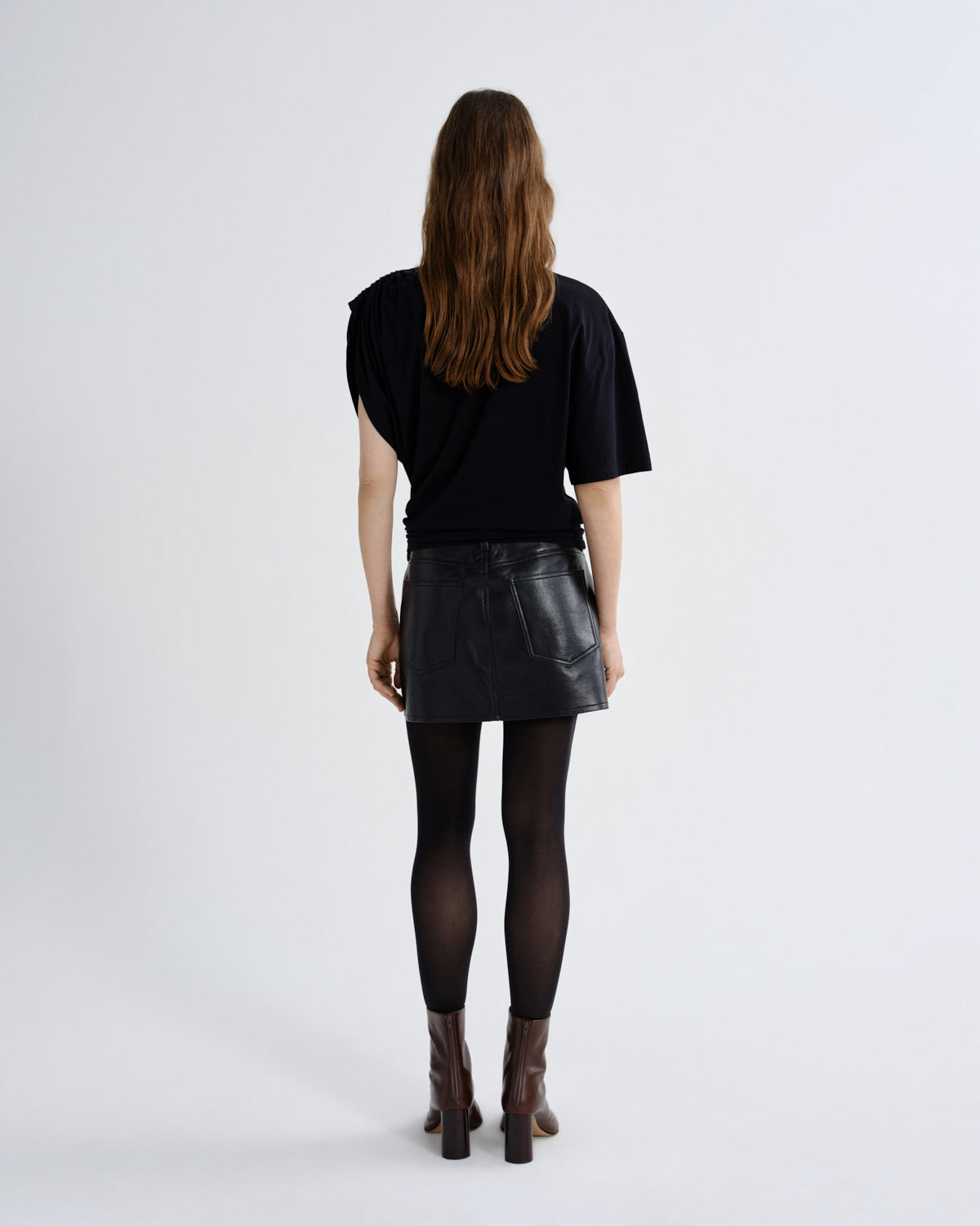 A model viewed from behind wearing a black short-sleeve top, a black faux-leather mini skirt, black tights, and brown heeled ankle boots against a plain light gray studio background.