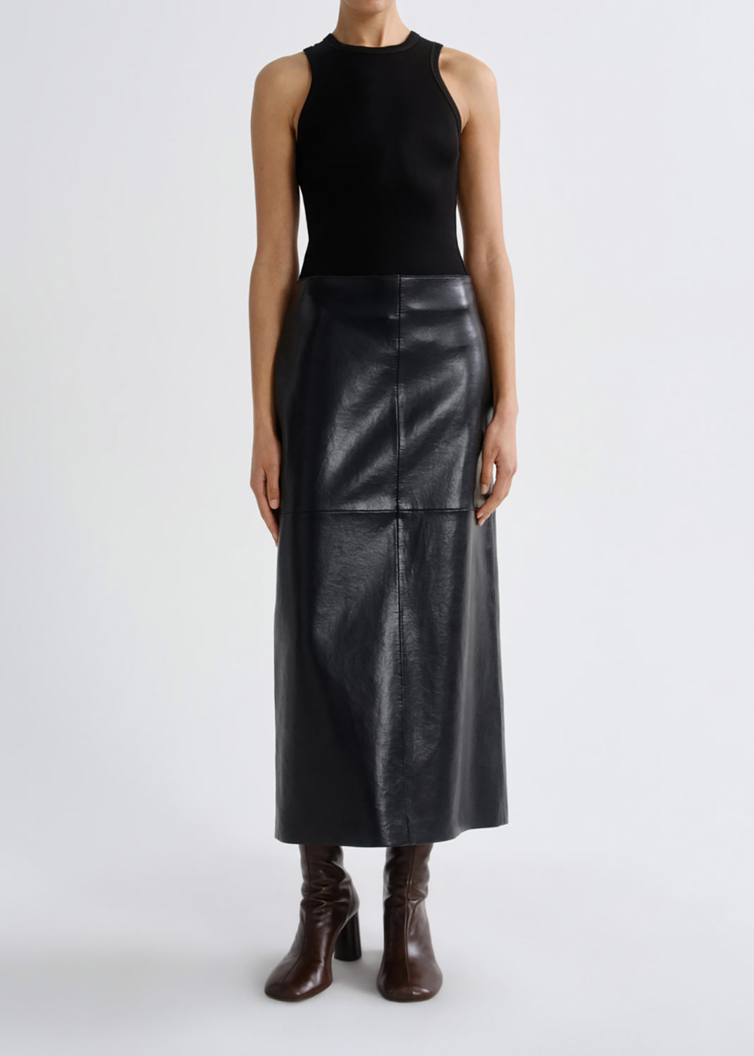 A model wearing a black sleeveless knit top and a black faux-leather midi skirt, paired with dark brown heeled ankle boots, against a grey background.