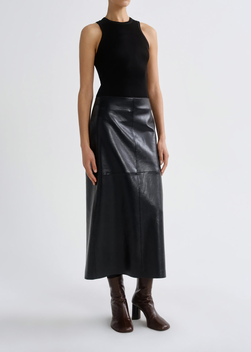 A model wearing a black sleeveless mock-neck knit top and a black faux-leather midi skirt, paired with dark brown heeled ankle boots, against a grey background.