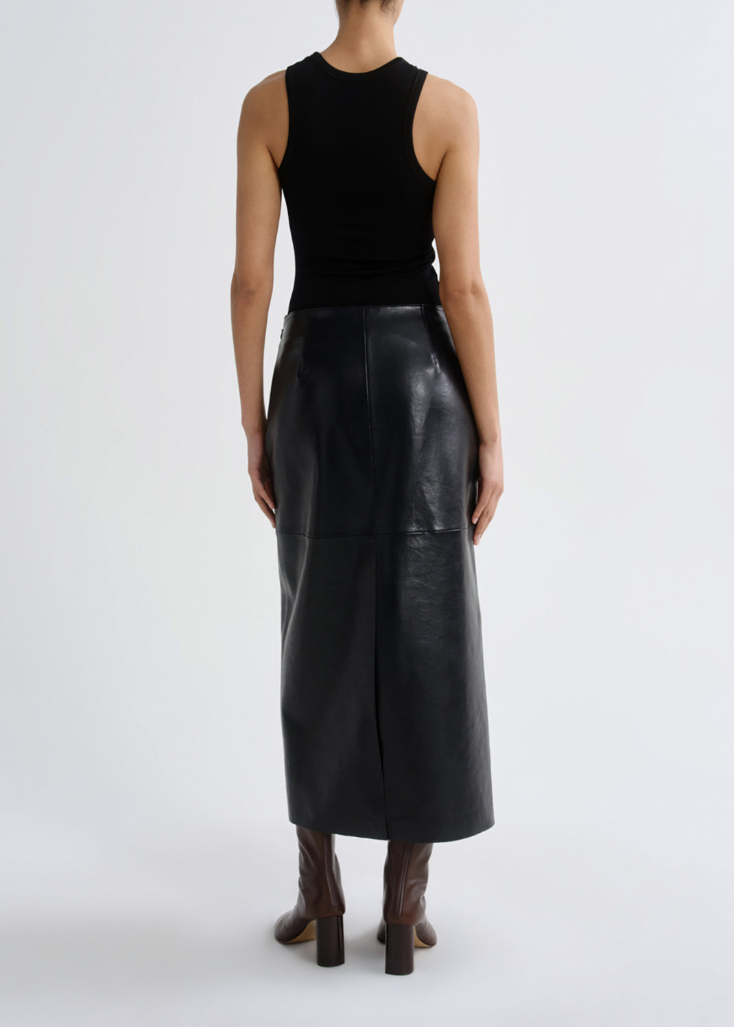 A model shown from the back wearing a black sleeveless ribbed top and a black leather midi skirt, paired with brown heeled boots, against a grey background.