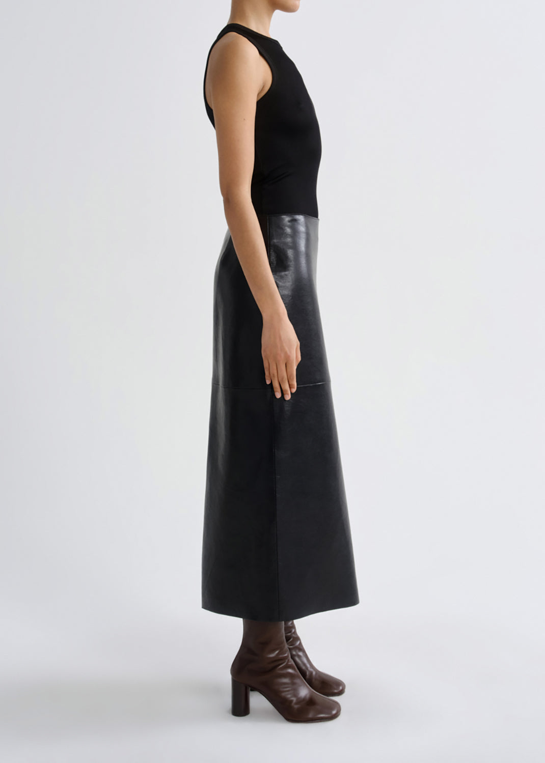 A model shown in profile wearing a sleeveless black ribbed knit top and a black leather midi skirt with a glossy panel, paired with dark brown heeled ankle boots, against a grey background.