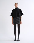 A model stands against a plain white studio background wearing a black short-sleeve mock-neck cape top, a black textured mini skirt, black tights, and black ankle boots.