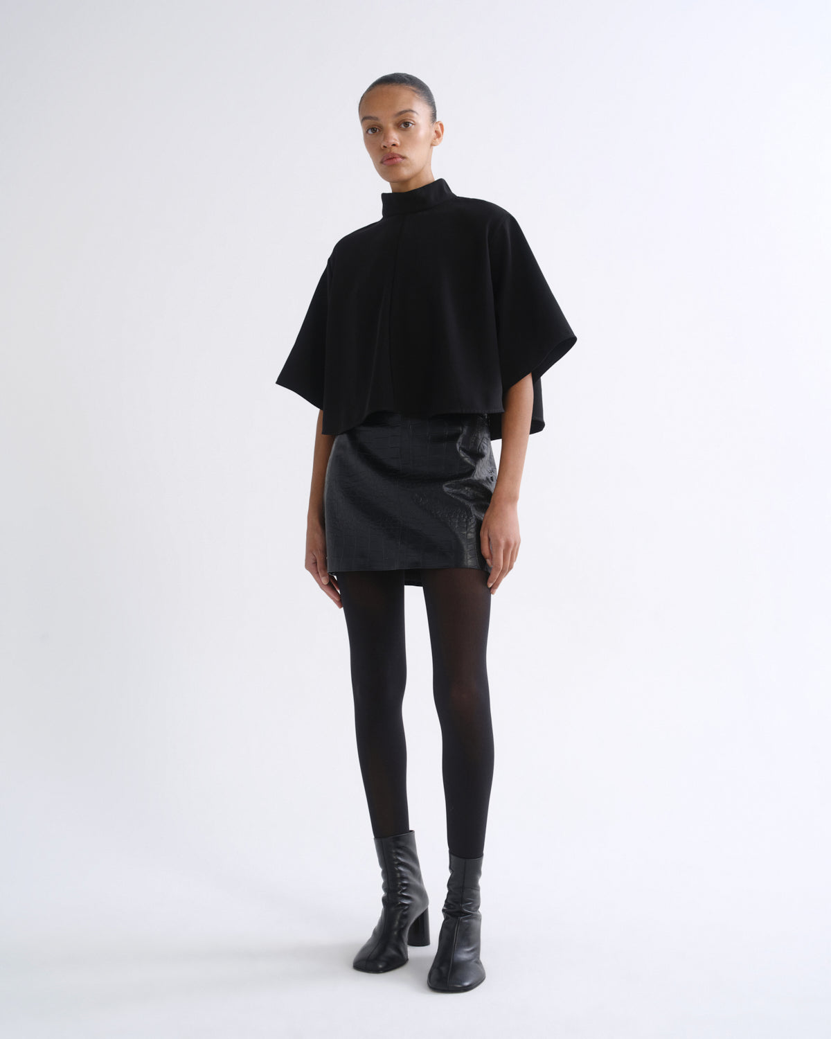 A model stands against a plain white studio background wearing a black high-neck cape-sleeve top, a black mini skirt with tonal side panels, black tights, and black ankle boots.