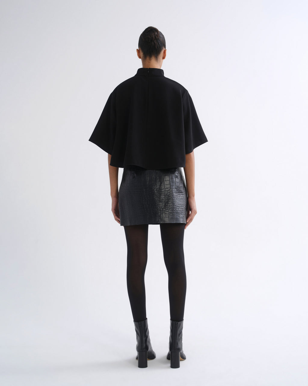 A model viewed from the back wearing a black short-sleeved high-neck top, a textured black mini skirt, black tights, and black ankle boots, set against a plain light gray studio background.