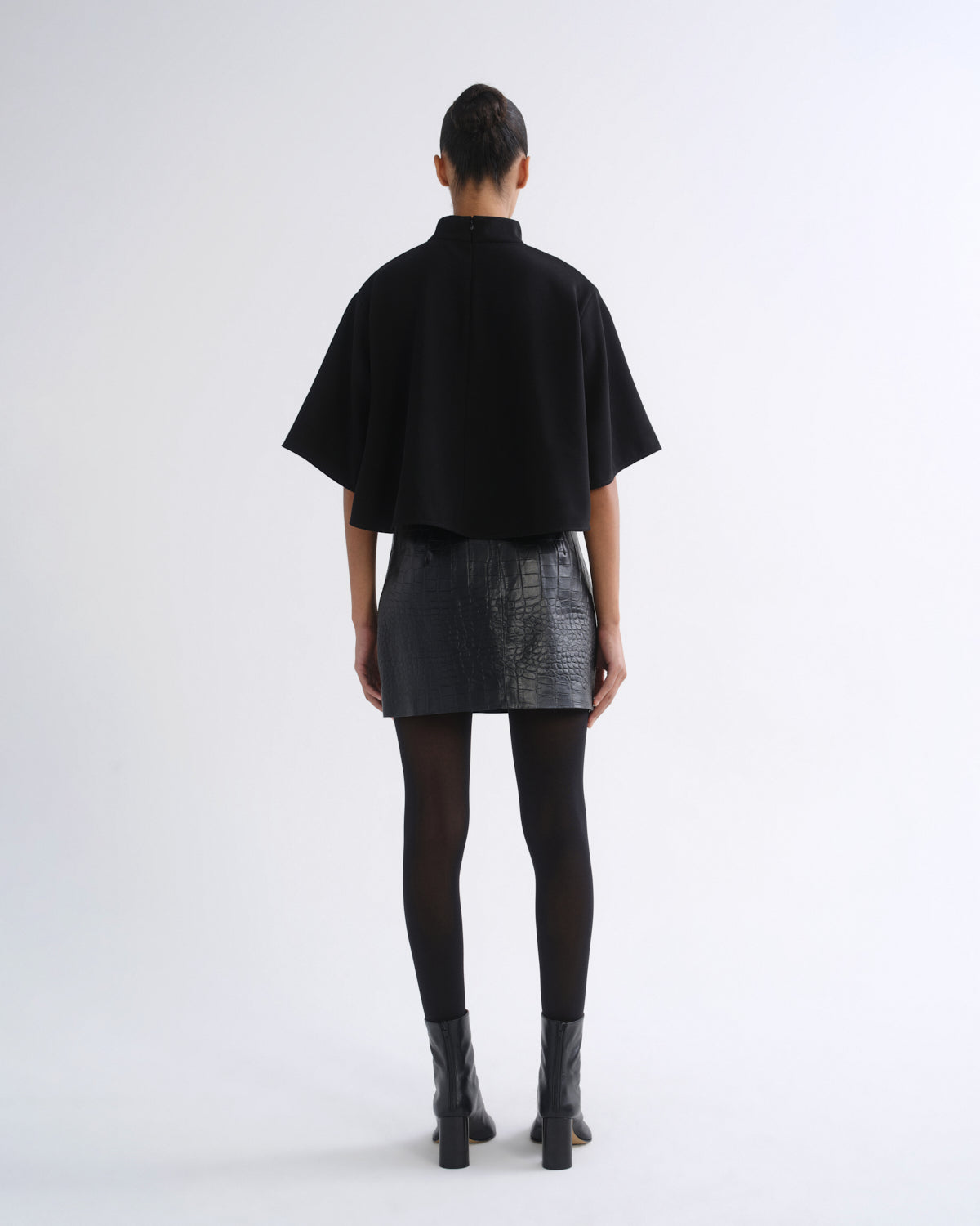A model viewed from the back wearing a black short-sleeved high-neck top, a textured black mini skirt, black tights, and black ankle boots, set against a plain light gray studio background.