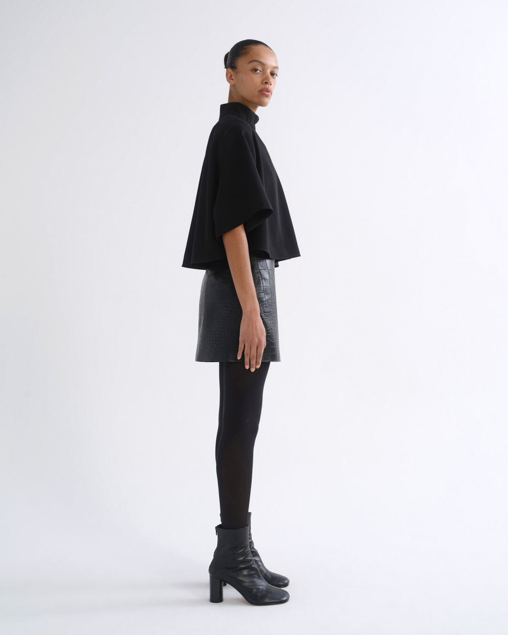 A model is shown in profile wearing a black cropped cape-sleeve top, a black faux-leather mini skirt, black tights, and black heeled ankle boots against a plain white studio background.