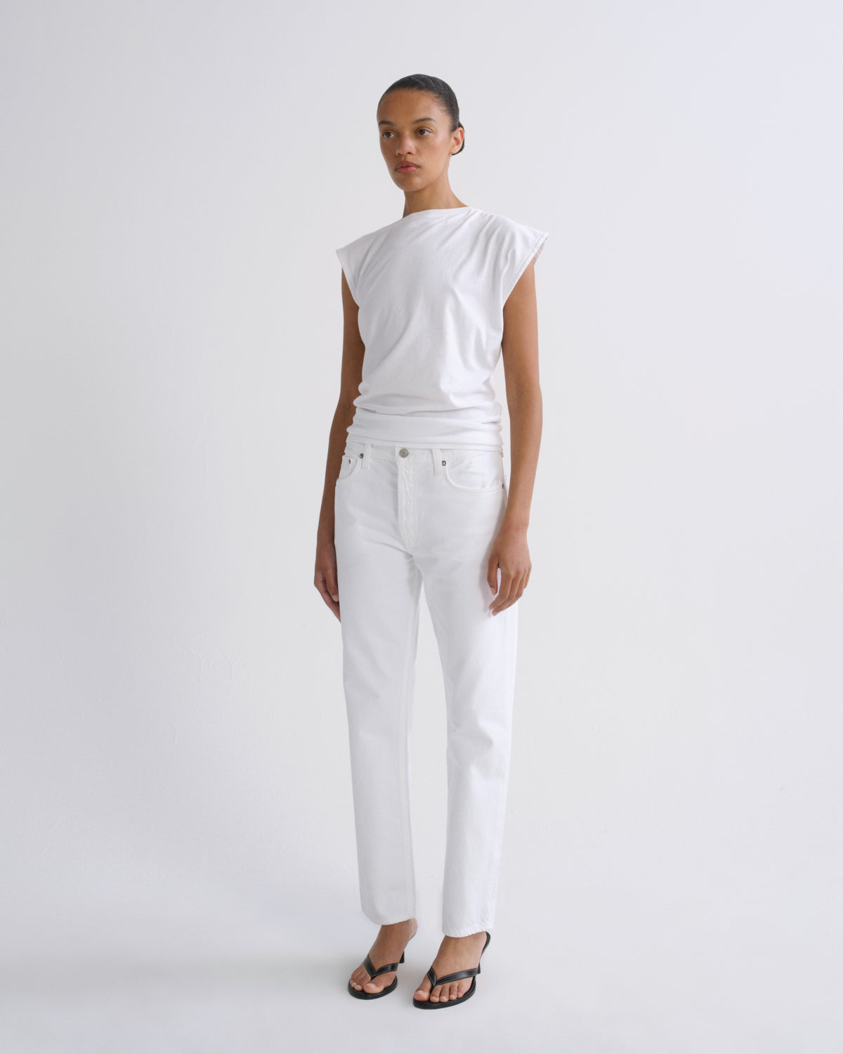 A model stands against a light gray studio background wearing a white sleeveless top, white straight-leg jeans, and black sandals.