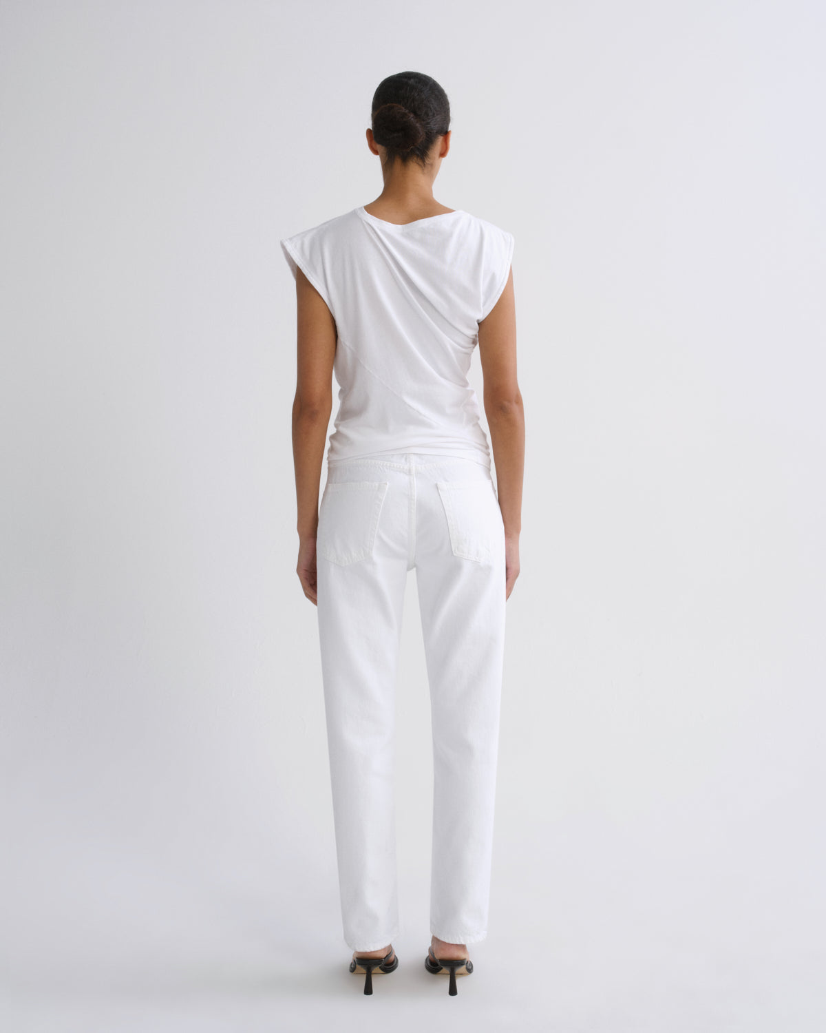 A model viewed from the back wearing a white draped sleeveless top and white trousers, standing against a light gray studio background.