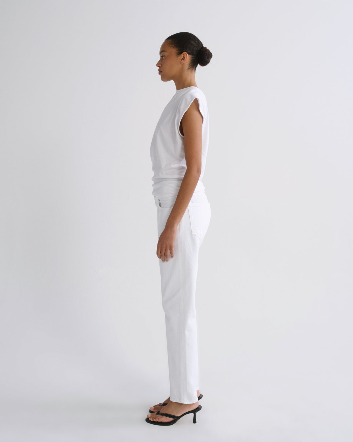 A model in profile wearing a white sleeveless top and white trousers, paired with black heeled sandals, standing against a plain white studio background.