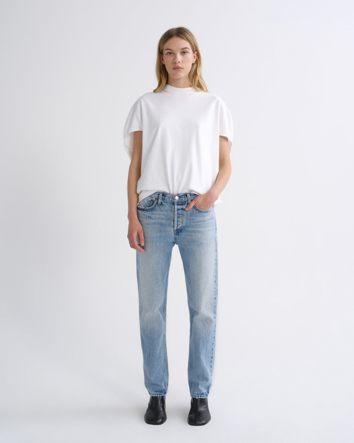 A model stands against a light gray studio background wearing a white short-sleeve t-shirt, light-wash blue jeans, and black boots.