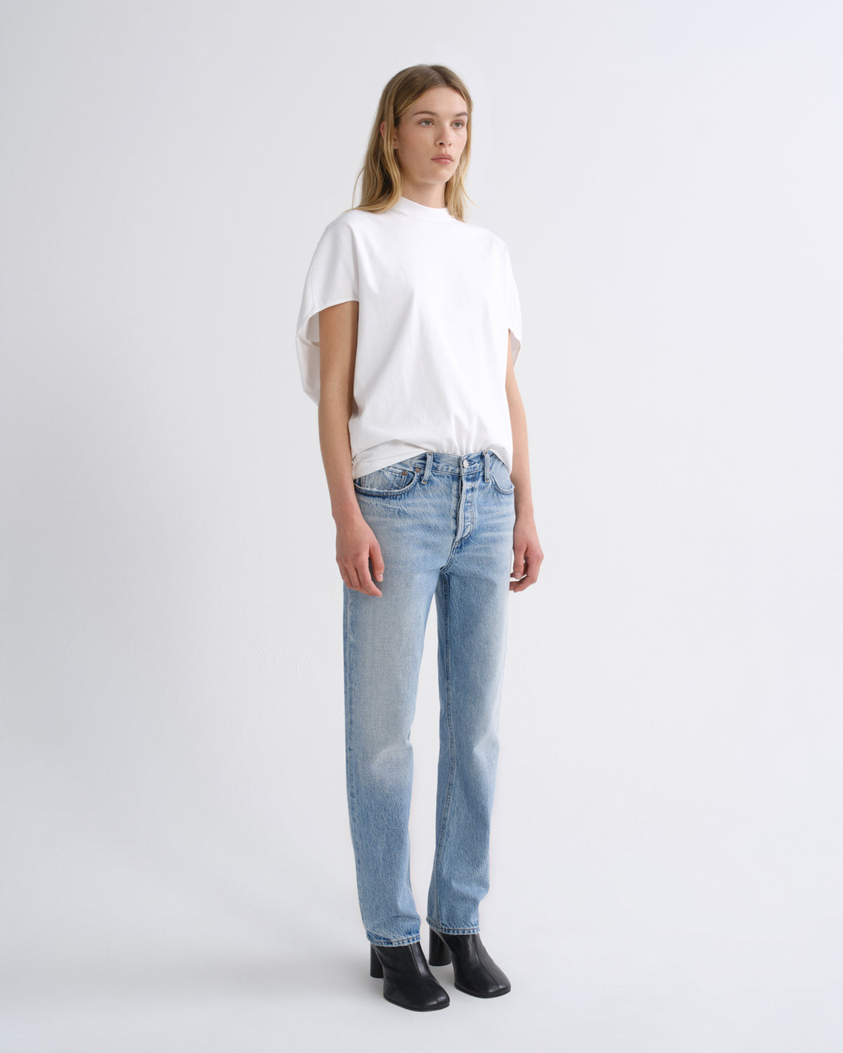 A model stands in a studio wearing a white short-sleeve mock-neck t-shirt, light-wash blue jeans, and black ankle boots against a light gray background.